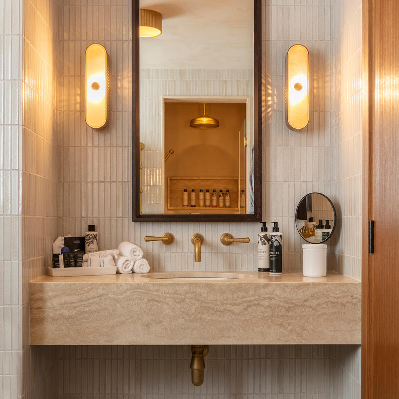 A beige marble sink sits under a framed mirror, flanked by two lights, in a bathroom with white tiled walls and a dark green tiled floor. Various toiletries and folded towels are arranged neatly on the countertop. A reflection in the mirror shows a partly