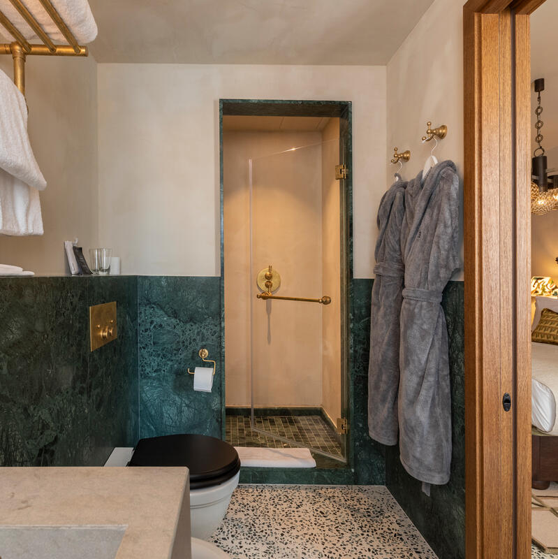 Bathroom with dark green marble walls and modern fixtures; includes a glass shower, black toilet seat, towel rack with white towels, and bathrobes hanging on hooks. Adjacent bedroom visible.