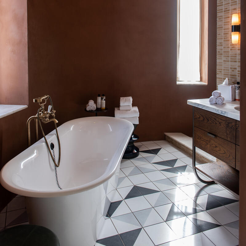 A white freestanding bathtub with bronze fixtures sits in a bathroom with brown walls, a patterned tile floor, and a tall window. A wicker light fixture hangs overhead.