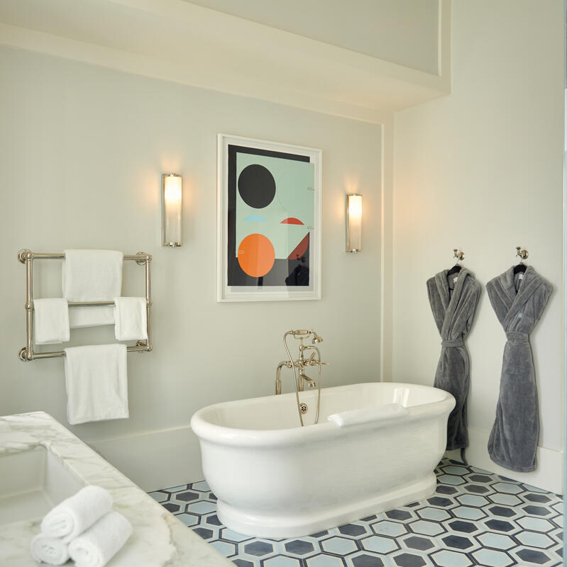 Bathtub surrounded by modern decor in a bright, elegant bathroom, featuring a towel rack, a framed abstract artwork, two gray robes on hooks, and geometric-patterned floor tiles.