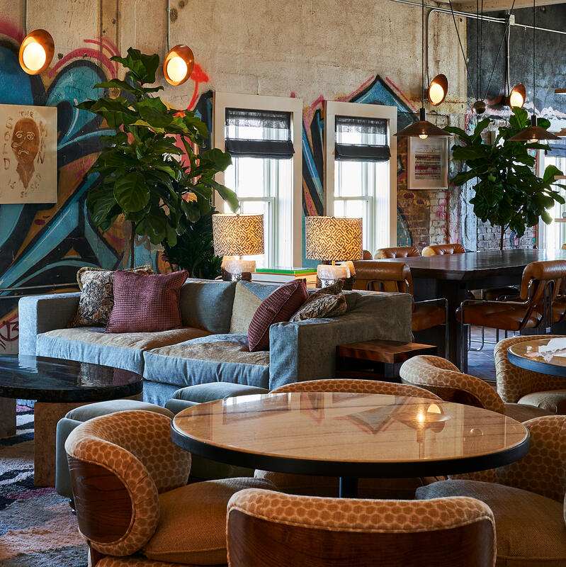 A modern lounge features a grey sectional sofa with purple cushions, surrounded by patterned armchairs and round tables. Graffiti-covered walls, large windows, plants, and eclectic lamps complete the vibrant setting.