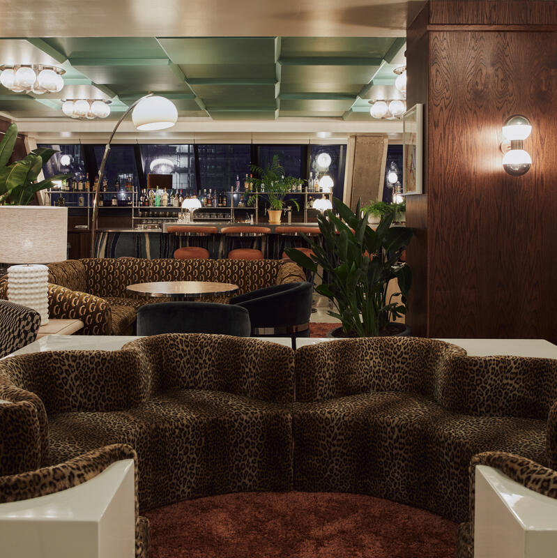 Leopard-print sofa sits in a stylish, dimly lit lounge with patterned chairs, surrounding tables, plants, retro light fixtures, wood accents, and a mirrored wall, leading to a well-stocked bar area.