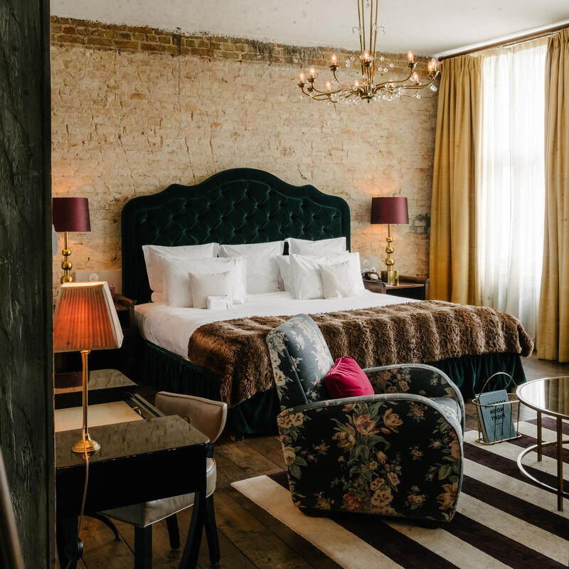 Luxurious bed with white pillows and brown throw blanket, positioned against a brick wall beneath a chandelier. Beside it is a floral armchair, gold lamp, and large windows with yellow curtains.