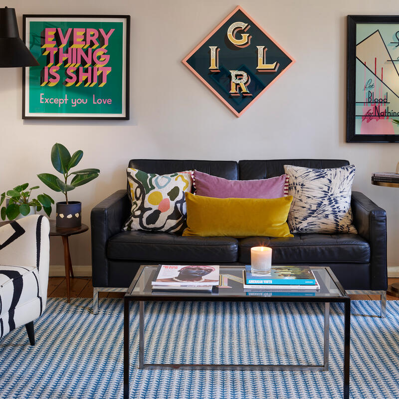 A stylish living room features a black sofa with colorful pillows, framed by modern wall art. A patterned armchair and a candle on a glass coffee table add warmth. Text includes "EVERYTHING IS SHIT Except you Love" and "Shave for a Penny."