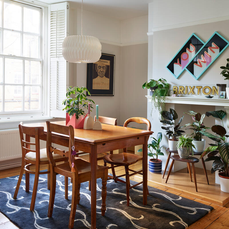 A wooden dining table with mismatched chairs under a white pendant lamp and surrounded by potted plants, located in a bright, homey room with a large window and decorative wall art reading "YOU WIN" and "BRIXTON."