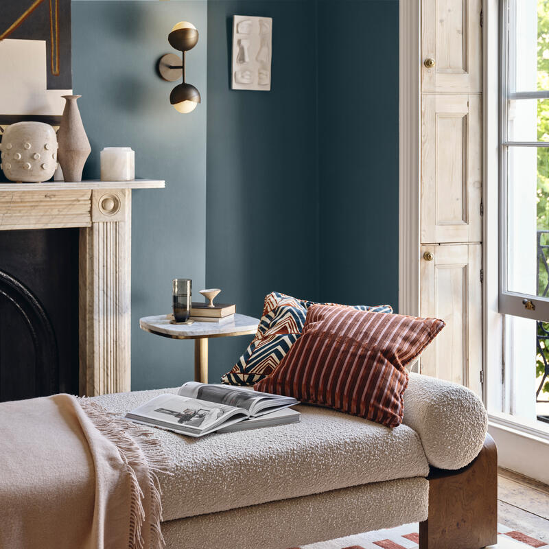 Beige daybed with patterned cushions and open magazine, placed in a cozy living room with blue walls, a marble fireplace adorned with vases and artwork, near a large window overlooking greenery.