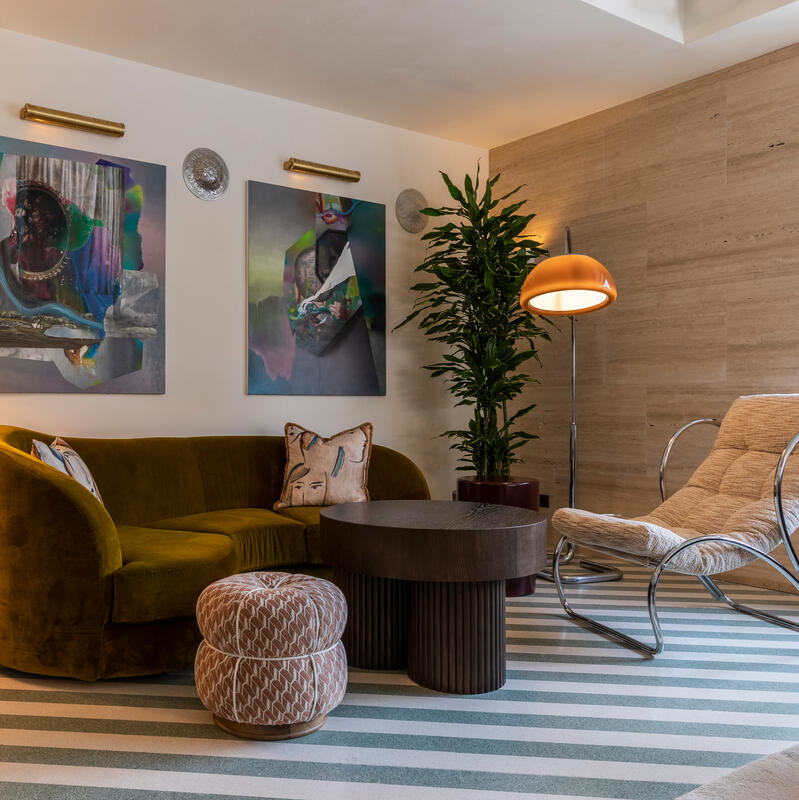 A living room features a green velvet sofa with cushions, a round coffee table, a modern lamp, an upholstered armchair, and two abstract paintings on the wall.