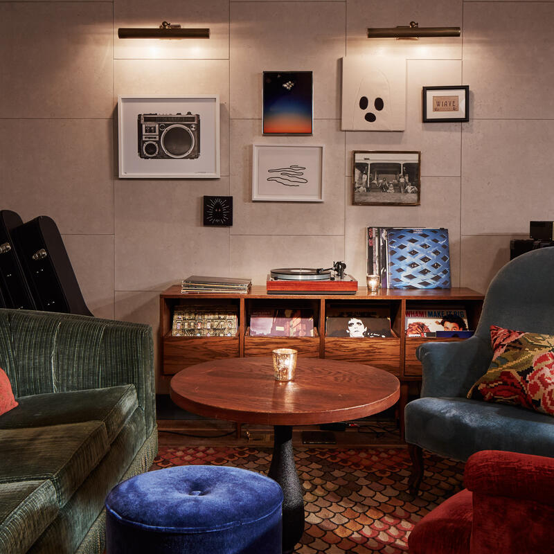 A cozy living room features a record player on a wooden console surrounded by records, framed art on the wall, green and blue armchairs, a candlelit coffee table, and guitar cases.