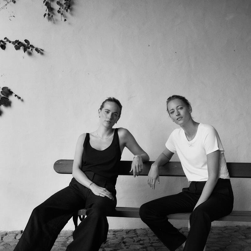 Two individuals sit casually on a bench against a textured wall with some hanging vines. One wears a black top and pants, the other a white top and black pants.