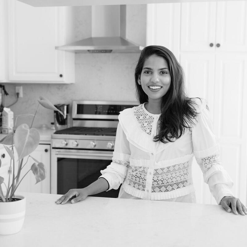 A woman, smiling, stands behind a countertop in a modern kitchen with white cabinets. A potted plant is on the countertop, and appliances are visible in the background.