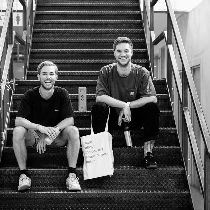 Two men sit on industrial staircase steps, smiling. One holds a beverage cup, the other holds a tote bag reading, "care about the ocean? show me your bottle." Indoor environment with signage.