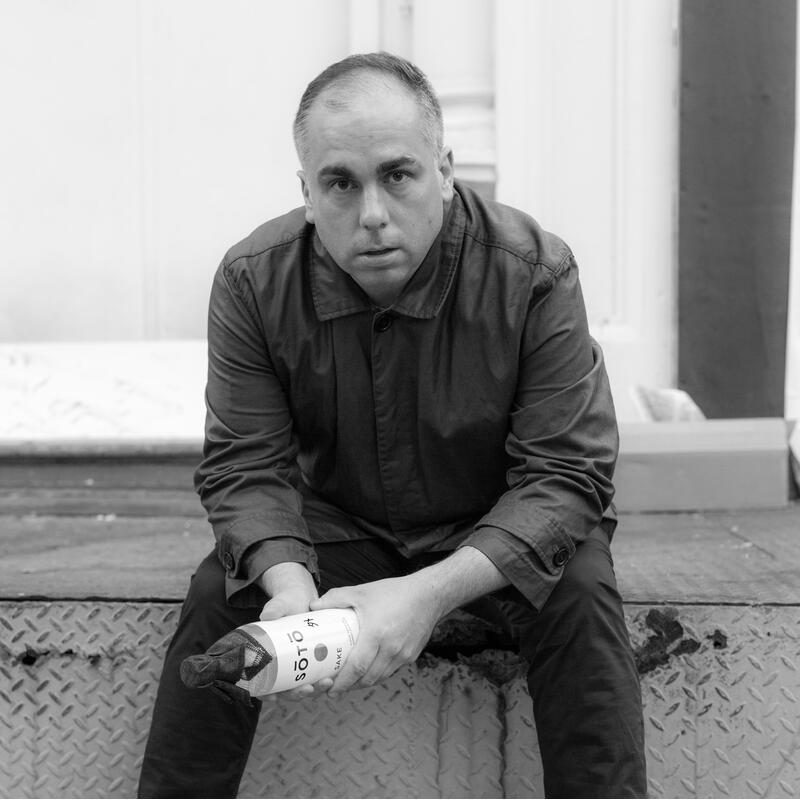 A person sits on a metal step while holding a bottle labeled "Solo Sake" in a partially wrapped cloth bag, set against an urban backdrop with visible concrete and metal textures.