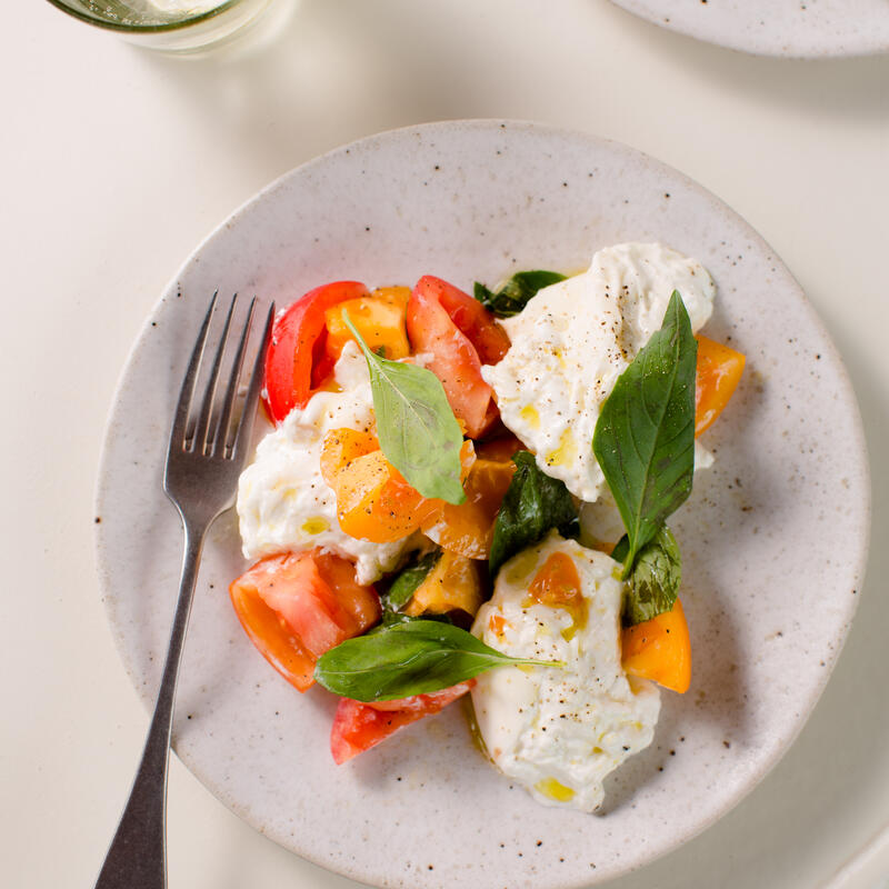 A plate of colorful tomato and cheese salad garnished with basil alongside a glass of lemon water, placed on a white table.