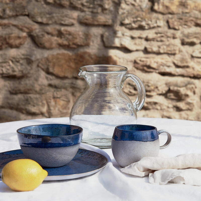 Glass pitcher, ceramic bowl, plate, and mug, and a lemon rest on a white tablecloth. They sit outdoors in front of a textured stone wall.