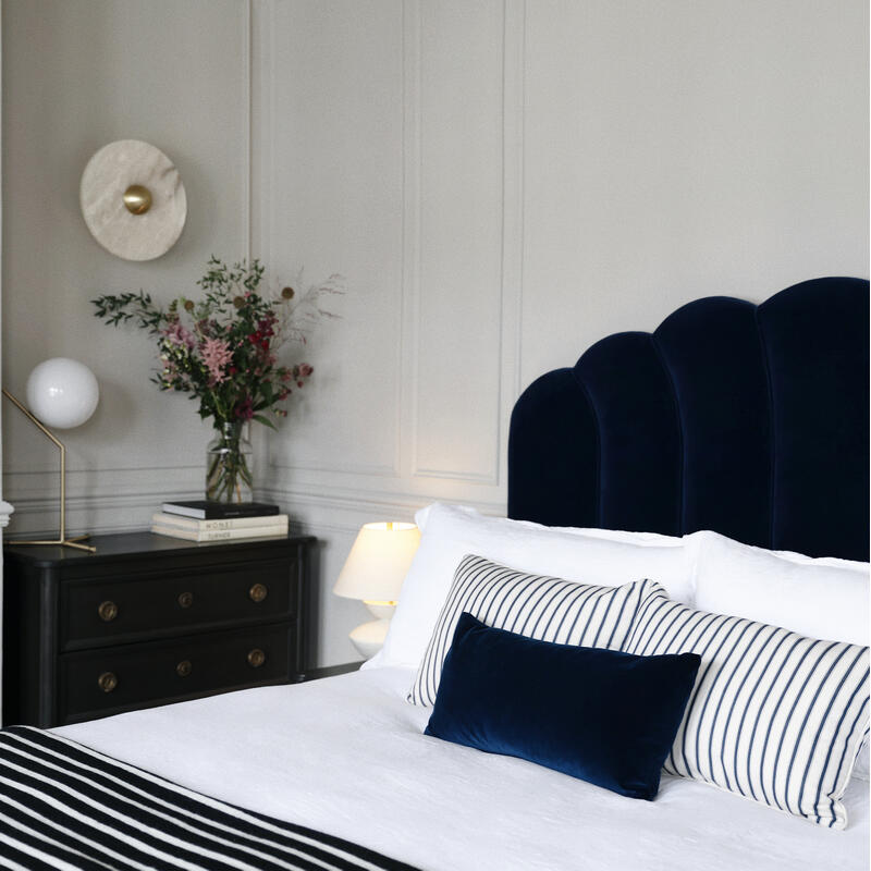 Bed with blue velvet headboard and striped pillows in a stylish room. Beside it, a black chest with books, flowers, and a lamp against a light grey wall with minimal decor.