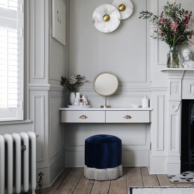 Vanity table with toiletries and a round mirror, accompanied by a blue stool placed by a window in a well-decorated room; a mantel above a fireplace holds flowers, a bust, and decor.