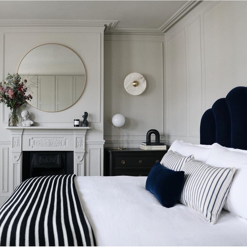 A bedroom features a bed with white linens and blue-striped pillows, adjacent to a fireplace with floral decor and a round mirror, nestled in an elegantly paneled, light-gray room.